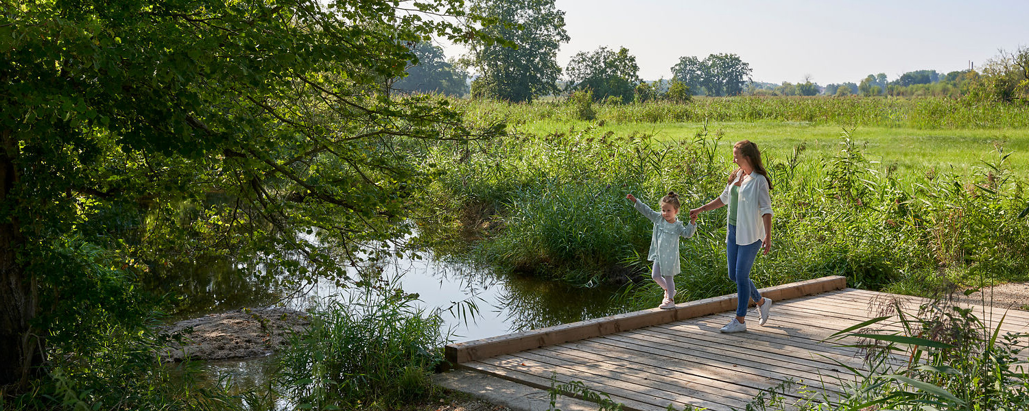 Eine Frau mit einem Kind an der Hand laufen über eine Brücke über einen Bach, umgeben von grünen  Wiesen.